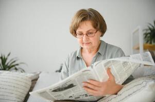 Senior senhora lendo dela jornal às casa relaxante em uma sofá e encarando sobre a topo às a espectador foto