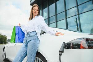 feliz jovem adulto mulher sorridente largo, olhando ausente, cobrando automóvel bateria a partir de pequeno público estação, em pé perto elétrico carro. foto