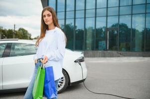 feliz jovem mulher em pé em cidade estacionamento perto elétrico carro, cobrando automóvel bateria a partir de pequeno cidade estação, segurando compras bolsas. foto