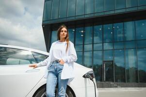 feliz jovem adulto mulher sorridente largo, olhando ausente, cobrando automóvel bateria a partir de pequeno público estação, em pé perto elétrico carro. foto