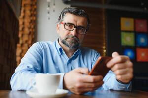 tendo tempo para pausa para o café. homem maduro confiante em trajes formais, bebendo café e digitando uma mensagem no celular enquanto está sentado no restaurante foto