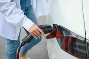 feliz jovem adulto mulher sorridente largo, olhando ausente, cobrando automóvel bateria a partir de pequeno público estação, em pé perto elétrico carro. foto