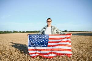 jovem patriótico agricultor carrinhos entre Novo colheita. Garoto caminhando com a americano bandeira em a trigo campo a comemorar nacional independência dia. 4º do Julho conceito. foto