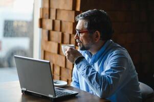 positivo Senior barbudo homem com cinzento cabelo bebendo café e usando computador portátil às cafeteria, cópia de espaço. à moda envelhecido homem de negocios dentro Borgonha Jaqueta desfrutando dele chá enquanto trabalhando on-line. foto