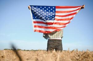 jovem patriótico agricultor carrinhos entre Novo colheita. Garoto caminhando com a americano bandeira em a trigo campo a comemorar nacional independência dia. 4º do Julho conceito. foto