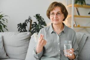 idoso mulher sentado em a sofá e levando pílulas às lar. pensionista com medicamentos foto