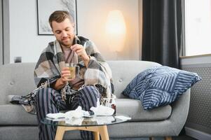 uma doente homem senta às casa em uma cinzento sofá com uma cobertor. doença, proteção, coronavírus, doença, gripe foto