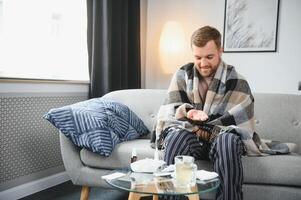 uma doente homem senta às casa em uma cinzento sofá com uma cobertor. doença, proteção, coronavírus, doença, gripe foto