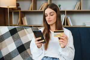 feliz adolescente fazendo compras conectados sentado em sofá às lar. jovem mulher inserir crédito cartão número em local na rede Internet com dela inteligente telefone. foto