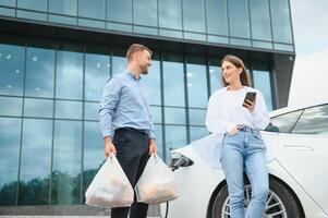 sorridente homem e mulher em a cobrando estação para elétrico carros. uma homem é cobrando uma carro, uma mulher é em pé com uma telefone foto