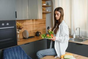 feliz sorridente fofa mulher é preparando uma fresco saudável vegano salada com muitos legumes dentro a cozinha às casa e tentando uma Novo receita foto