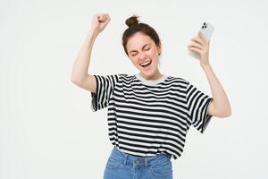 menina dançando com Móvel telefone contra branco fundo. mulher com Smartphone cantando, levantando mãos acima e a comemorar foto