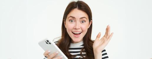 retrato do feliz, surpreso jovem mulher, segurando Móvel telefone, mostrando espantado reação para ótimo notícias, em pé com Smartphone sobre branco fundo foto