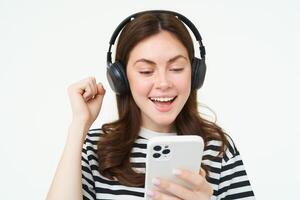 alegre, lindo jovem mulher usando Smartphone aplicativo, a comemorar, olhando feliz enquanto jogando com Móvel telefone, ouvindo música dentro sem fio fones de ouvido foto