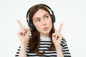 retrato do jovem morena mulher dentro fones de ouvido, ouvindo música dentro fones de ouvido, apontando dedos acima, mostrando bandeira, anúncio, isolado sobre branco fundo foto