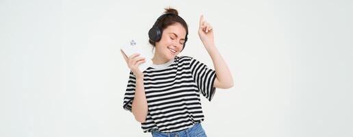 retrato do feliz mulher com Smartphone alterar música em Móvel telefone transmissão aplicativo, escuta música dentro fones de ouvido, dançando contra branco fundo foto