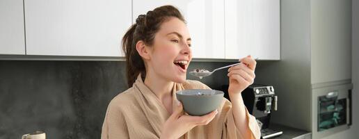 linda mulher começa dela dia com cereais, come café da manhã e sorrisos, desgasta roupão de banho, inclina-se em cozinha bancada, começa dela manhã com rápido refeição foto