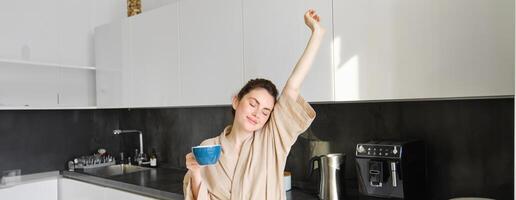 retrato do feliz menina dançando com café dentro a cozinha, vestindo roupão de banho, desfrutando dela manhã rotina foto