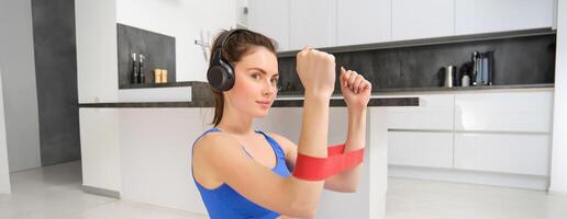 jovem mulher fazendo ginástica exercite-se às lar, usando elástico resistência banda em braços, músculo exercícios, sentado em borracha esteira dentro vivo quarto foto