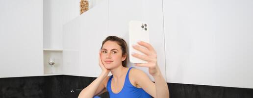 retrato do ginástica menina posando para foto, levando selfie em Smartphone aplicativo, sentado dentro cozinha, vestindo roupa ativa foto