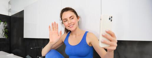 retrato do jovem ginástica garota, social meios de comunicação blogueiro, viver transmissão, dizendo Olá às Smartphone, acenando mão e sorridente, vestindo roupa ativa foto