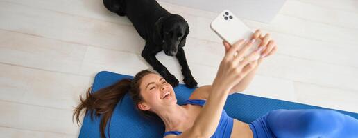 retrato do feliz jovem ginástica mulher, deitado em esteira e levando selfie com uma cachorro dentro vivo sala, exercite-se com dela animal foto