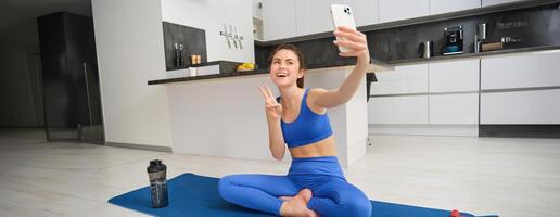 ativo jovem mulher, vlogger faz Esportes, registros dela exercite-se Treinamento a partir de casa em Smartphone Câmera, posando para selfie dentro dela casa, senta em borracha ioga esteira dentro azul perneiras e sportsbra foto