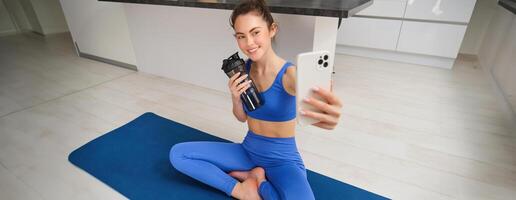 retrato do mulher levando selfie com água garrafa, ginástica instrutor mostra dela exercícios, fazendo exercite-se a partir de lar, em borracha azul ioga esteira, vestindo roupa de esporte foto