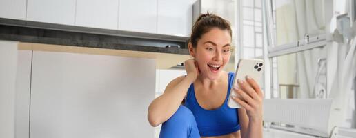 fechar acima tiro do jovem feliz mulher, fazendo Esportes, exercite-se às lar, verificação dela telefone, olhando espantado às Smartphone tela foto