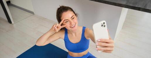 retrato do lindo fêmea atleta, mulher fazendo exercite-se às casa em borracha ioga esteira, leva selfie em Smartphone, faz fotos para social meios de comunicação do ginástica instrutor