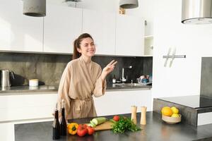 saudável estilo de vida. jovem mulher dentro roupão de banho preparando comida, cortar vegetais, cozinhando jantar em cozinha contador, em pé sobre branco fundo foto