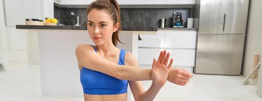 fechar acima tiro do mulher fazendo exercite-se a partir de lar, aquecer alongamento mãos, faz ginástica exercícios dentro azul sportsbra e sorridente foto