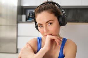 fechar acima retrato do morena ginástica garota, escuta música dentro sem fio fones de ouvido, sorrisos às Câmera, faz exercite-se sozinho às lar, senta em chão e exercícios foto