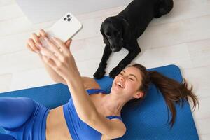 retrato do feliz jovem ginástica mulher, deitado em esteira e levando selfie com uma cachorro dentro vivo sala, exercite-se com dela animal foto