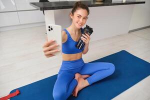 retrato do mulher levando selfie com água garrafa, ginástica instrutor mostra dela exercícios, fazendo exercite-se a partir de lar, em borracha azul ioga esteira, vestindo roupa de esporte foto