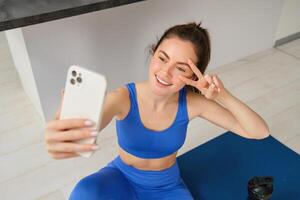 retrato do lindo fêmea atleta, mulher fazendo exercite-se às casa em borracha ioga esteira, leva selfie em Smartphone, faz fotos para social meios de comunicação do ginástica instrutor