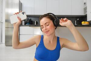 felicidade, Esportes e bem-estar. jovem mulher dançando dentro fones de ouvido, segurando Smartphone, fazendo exercite-se às lar, ginástica Treinamento dentro dela casa foto