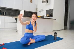 jovem sorridente mulher, aptidão menina fazendo exercite-se a partir de lar, senta em ioga esteira com Smartphone, levando selfie, registros esporte vlog dentro de casa foto