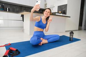 jovem sorridente mulher, aptidão menina fazendo exercite-se a partir de lar, senta em ioga esteira com Smartphone, levando selfie, registros esporte vlog dentro de casa foto