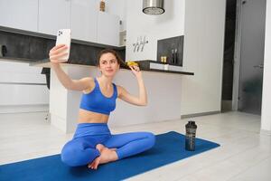 retrato do ativo garota, vlogger faz Esportes, registros dela exercite-se Treinamento a partir de casa em Smartphone Câmera, posando para selfie dentro dela casa, senta em borracha ioga esteira dentro azul perneiras e sportsbra foto