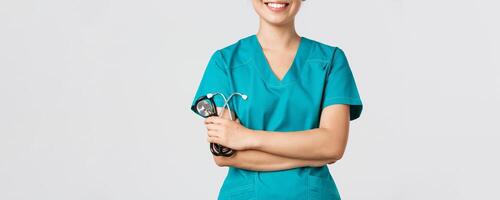 COVID-19, coronavírus doença, cuidados de saúde trabalhadores conceito. cortada tiro do ásia fêmea médico corpo sorridente, Cruz braços peito confiante e segurando estetoscópio, em pé branco fundo foto