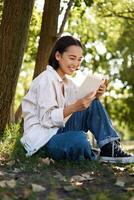 vertical tiro do feliz ásia mulher relaxante ao ar livre dentro parque, lendo dela livro e sentado debaixo árvore sombra em caloroso ensolarado dia foto