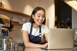 entusiasmado ásia barista, menina dentro avental, olhando às computador portátil, vídeo bate-papos, falando on-line, em pé atrás contador dentro café fazer compras foto