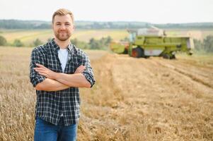 feliz agricultor orgulhosamente em pé dentro uma campo. combinar colheitadeira motorista indo para colheita rico trigo colheita. agrônomo vestindo flanela camisa, olhando às Câmera em uma terras agrícolas foto