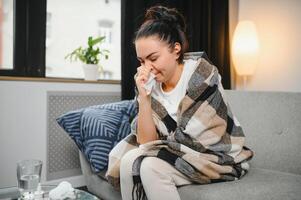 eu vou jovem mulher sentar em sofá coberto com cobertor, congelando, sopro dela escorrendo nariz, espirrar dentro tecido. fêmea obteve febre, gripe e gripe sintomas tosse às lar. doente alérgico com respiratório doença foto