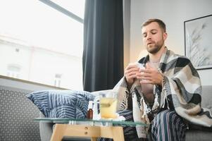 uma doente homem senta às casa em uma cinzento sofá com uma cobertor. doença, proteção, coronavírus, doença, gripe foto