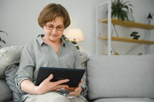 aposentadoria lazer. sorridente idosos mulher em repouso com digital tábua em sofá às lar, navegando Internet ou assistindo filmes foto