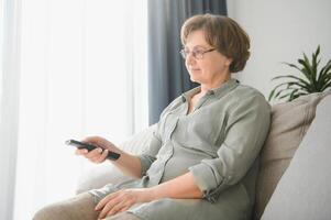 quebra notícias. chocado Senior mulher assistindo televisão às lar, sentado em sofá com controlo remoto controlador dentro mãos foto