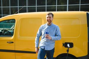 bonito homem bebendo café enquanto cobrando elétrico carro foto