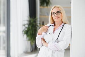 retrato do meio era fêmea médico é vestindo uma branco médico casaco com uma estetoscópio por aí dela pescoço. sorridente médico em pé às privado clínica foto